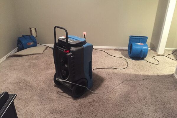 Carpet drying with dehumidifier and fans in a room, gray carpet, beige walls, cables on floor, equipment for flooding & water damage.