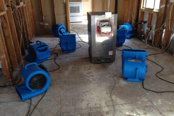 Blue industrial fans and dehumidifier drying a stripped room with water & flood damage.